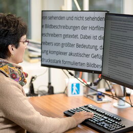 Eine Frau sitzt an einem Schreibtisch, vor sich zwei Bildschirme. Auf dem linken Bildschirm ist die Schrift sehr stark vergrößert.